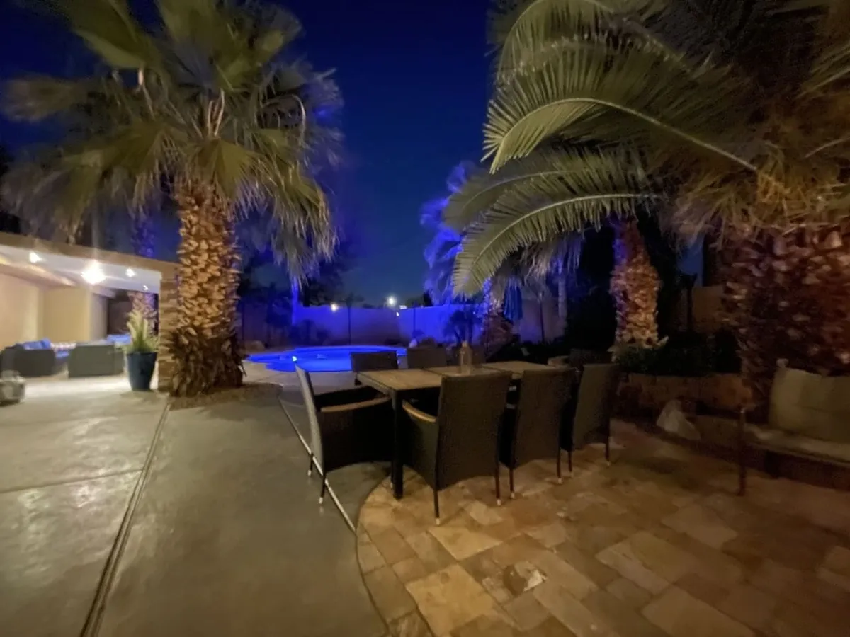 An elegant evening scene of a backyard oasis with palm trees, a beautifully set outdoor dining table, and a swimming pool illuminated with blue lighting. The mood is calm and inviting, ideal for stargazing and hosting gatherings under the night sky.