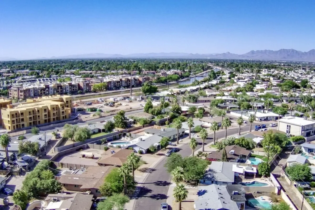 A scenic view of a winding river flanked by a vibrant cluster of residential houses, a modern new construction building, and the surrounding mountains. People can be seen strolling along the riverside, exploring nearby shops, and enjoying the outdoor ambiance of the area.