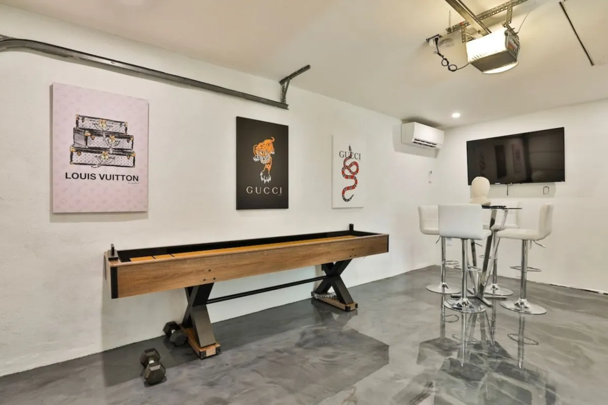 A stylish, upscale interior room featuring a shuffleboard table, luxury-brand artwork (Gucci and Louis Vuitton), a bar table with stools, and a TV. The décor is chic and playful, ideal for socializing or unwinding after a night out.
