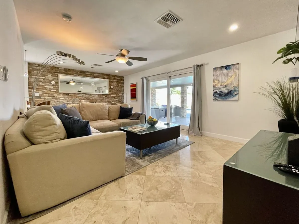 This interior features a cozy sectional sofa, a stylish coffee table, wall-mounted decorative art, and large sliding glass doors that allow sunlight to stream in. The arrangement creates a seamless flow between lounging and entertaining, reinforcing both relaxation and celebration.