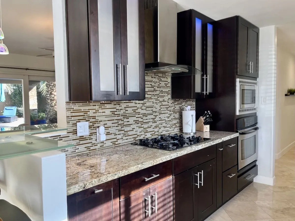 This image showcases a contemporary kitchen featuring granite countertops, stainless steel appliances, and a striking glass mosaic backsplash. The space is filled with natural light and arranged for both functionality and style, inviting guests to gather around for cooking and conversation.