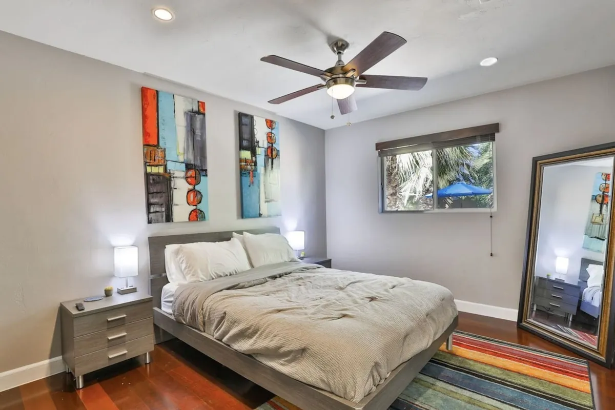 A serene bedroom with a plush bed adorned with soft linens, stylish wall art above the headboard, a window filtering in natural light, and a mirror adding depth to the tranquil space. The room exudes calm, inviting relaxation and rest.