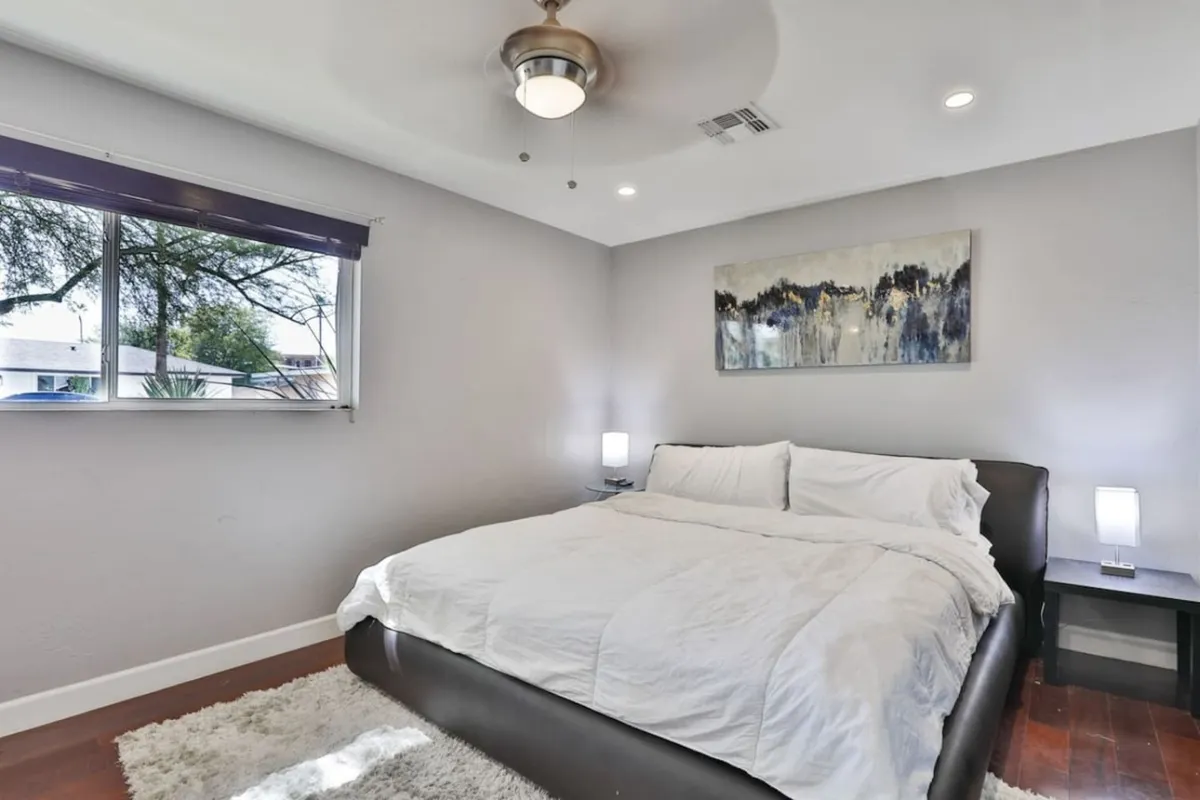 A tranquil bedroom space with a large bed dressed in crisp white linens, contemporary artwork, nightstands with soft lighting, and a window offering views and natural light. The setting is both inviting and peaceful, perfect for relaxation.