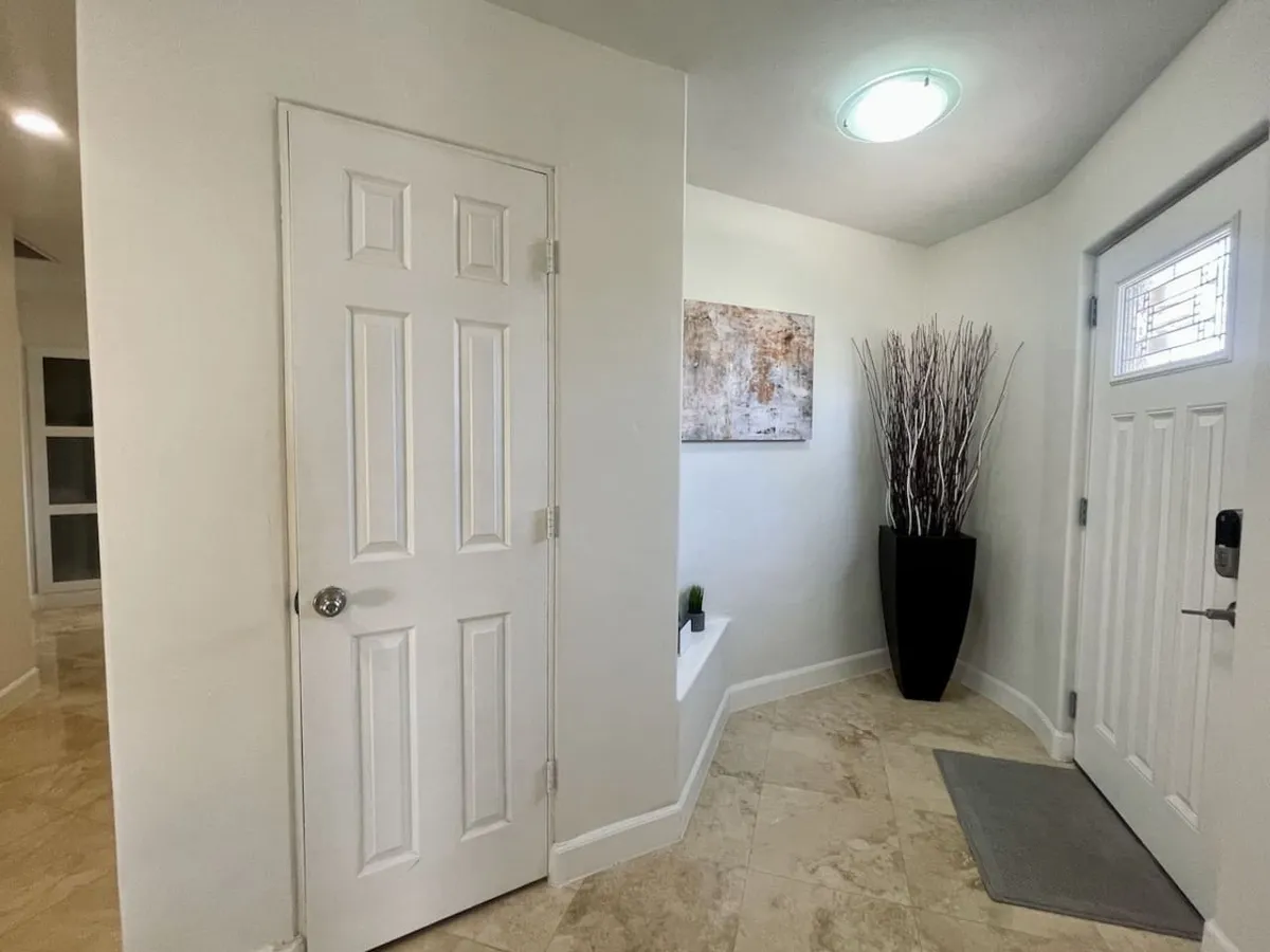 This image captures a welcoming entryway with a white door, decorative wall art, a tall plant in a vase, and an inviting entryway mat. The space suggests the first step into a stylish and comfortable home, balancing warmth and sophistication.