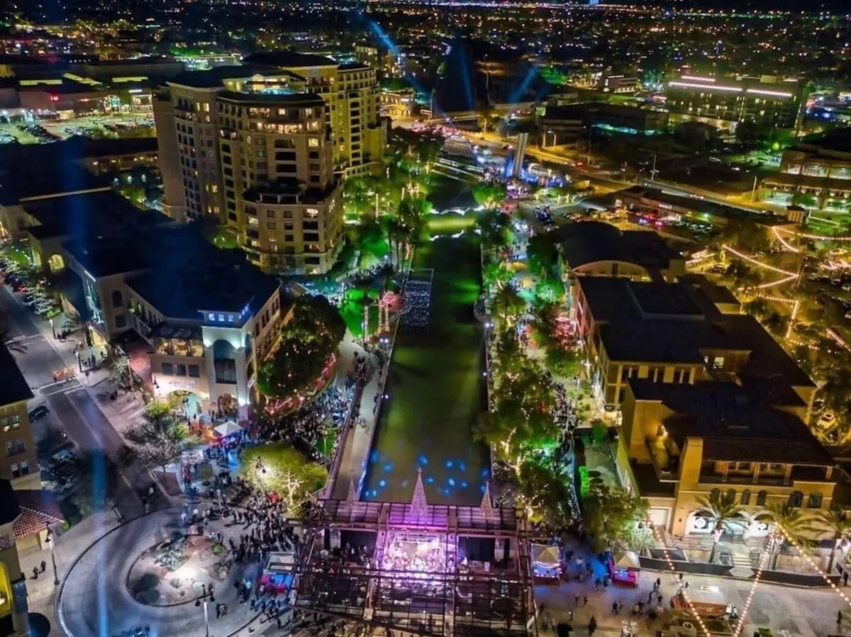A vibrant nighttime scene featuring illuminated buildings, a lively crowd, a water feature, and a stage hosting a performance. The area is energetic, evoking the festive and social atmosphere of a popular nightlife district.